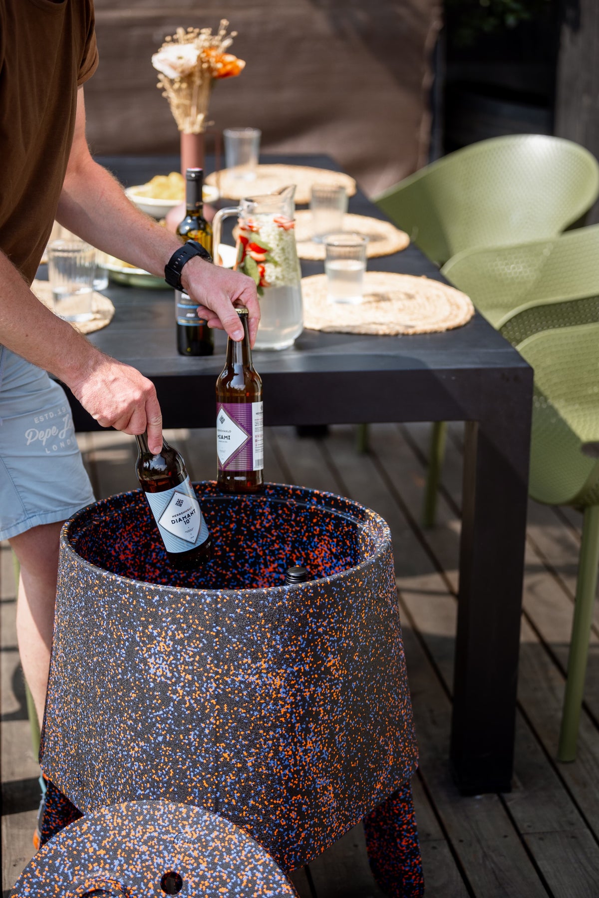 Hand taking a bottle out of AYTO Orange Mix used as a drinks cooler next to an outdoor dining table